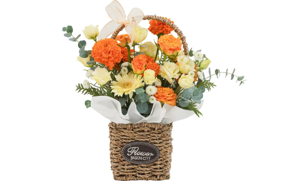 Yellow and orange flower basket with marigolds, roses and gerbera in a woven natural basket with a handle and sheer peach bow