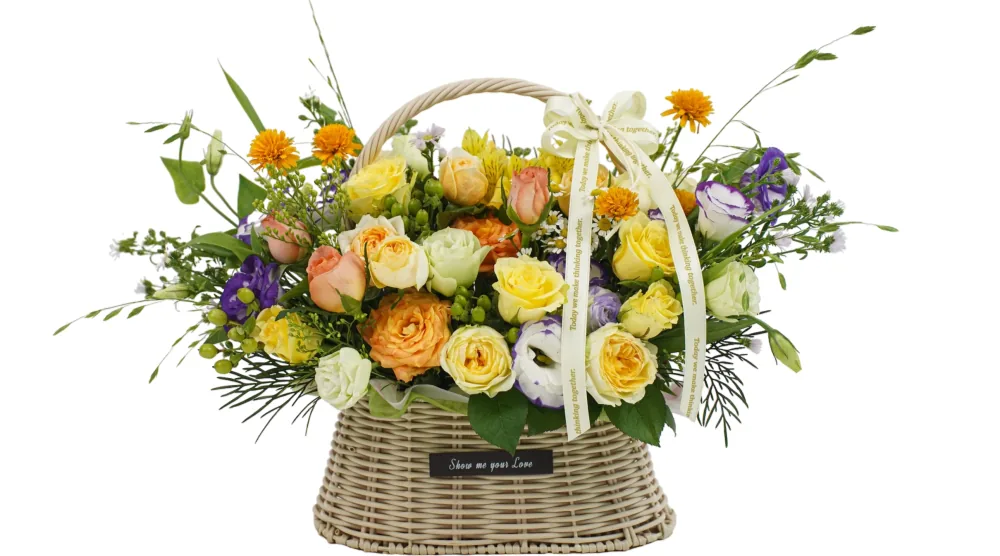 Wide flower basket with yellow and apricot roses, purple-edged lisianthus, orange pompoms and wild greenery in a pale wicker basket with a cream ribbon