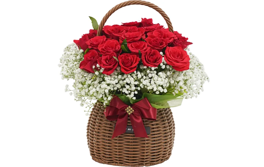 Red roses massed above white baby's breath in a handled wicker basket with a burgundy bow