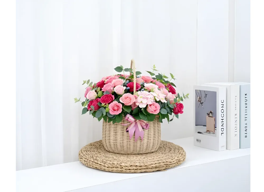 Giỏ hoa xà phòng hồng trên tấm thảm đan đặt trên kệ trắng cạnh sách, phía sau là rèm mỏng