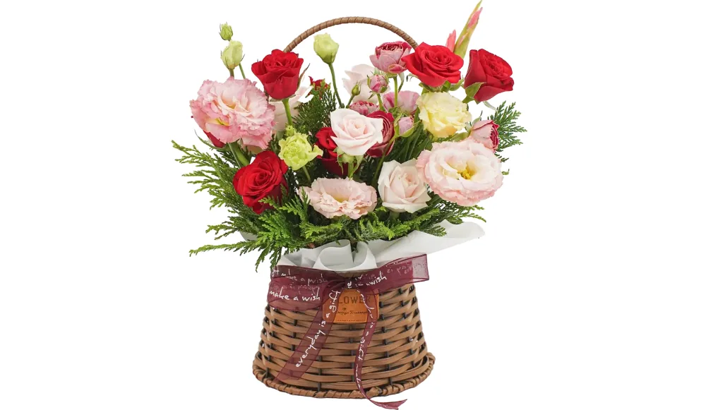 Red roses, peach and pale yellow lisianthus, spray roses and conifer greens in a brown wicker basket with a burgundy ribbon