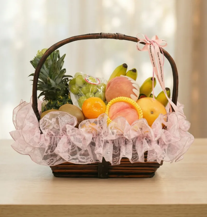 Giỏ mây sẫm màu có quai viền ren hồng và ruy băng hồng, đầy dứa, nho xanh, đào, chuối, kiwi và cam quýt