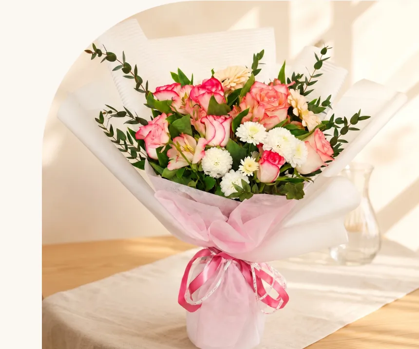 Bouquet of pink-edged cream roses, white button chrysanthemums and peach blooms with eucalyptus, wrapped in pink tulle and tied with a pink satin ribbon, standing on a wooden table